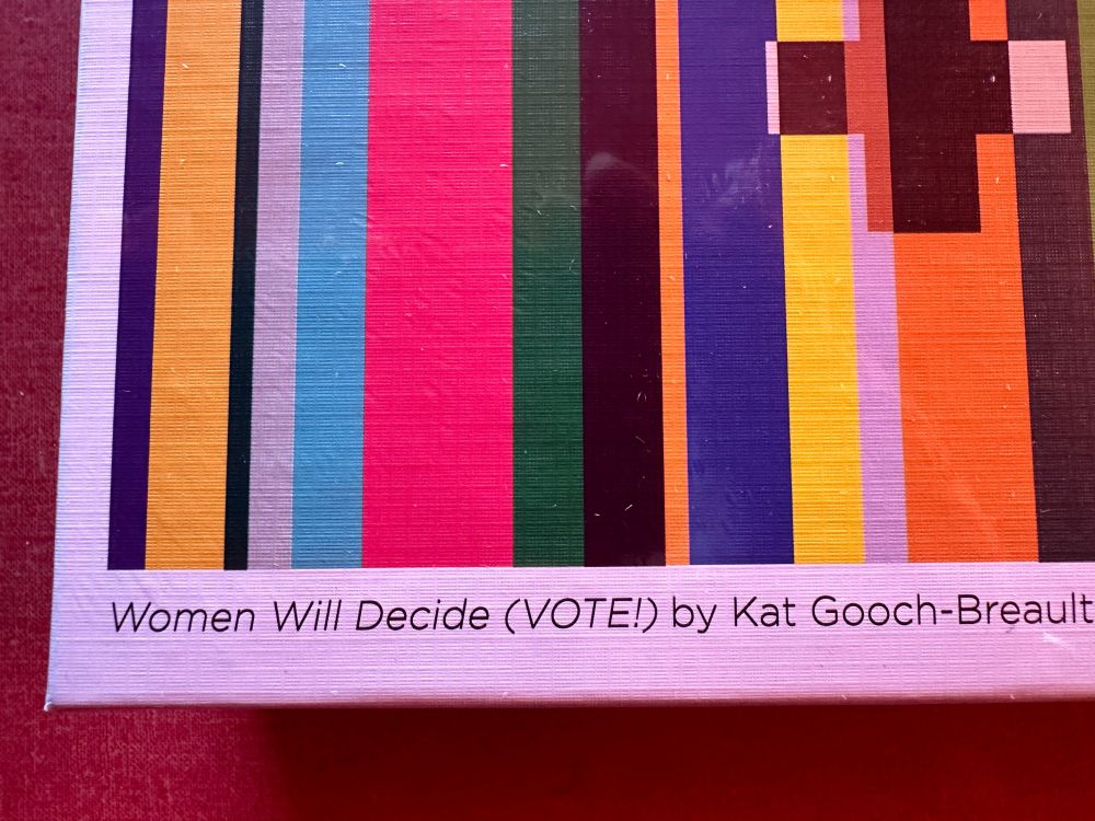 Close up of the credit/title of the colorful jigsaw puzzle.  The credit and title on lower left half of the puzzle box reads "Women Will Decide (VOTE!)" by Kat Gooch-Breaul.