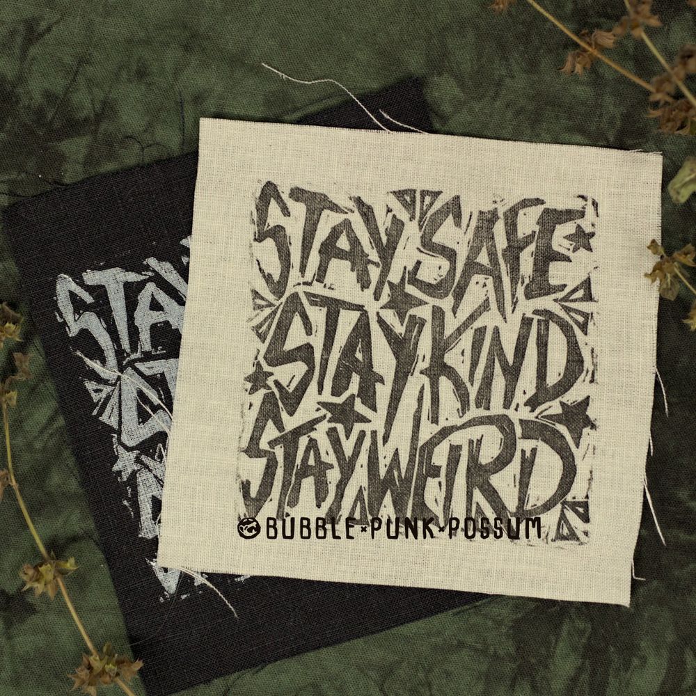 Two linen patches stacked on a green background. The top patch is white and the bottom one is black and they share the same design of the phrase "Stay Safe Stay Kind Stay Weird".