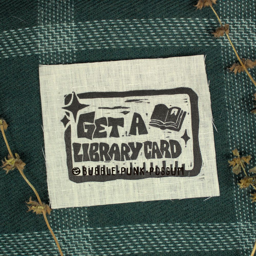 A white patch stacked on top of green flannel. The patch design features a rectangular border and the phrase "Get a Library Card" with an open book in the top right corner and sparkles in the top left corner. 