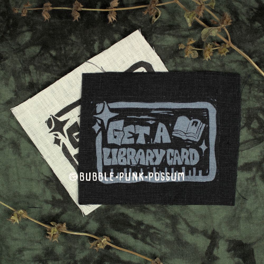 A black patch stacked on top of a white patch. The patch design features a rectangular border and the phrase "Get a Library Card" with an open book in the top right corner and sparkles in the top left corner. 