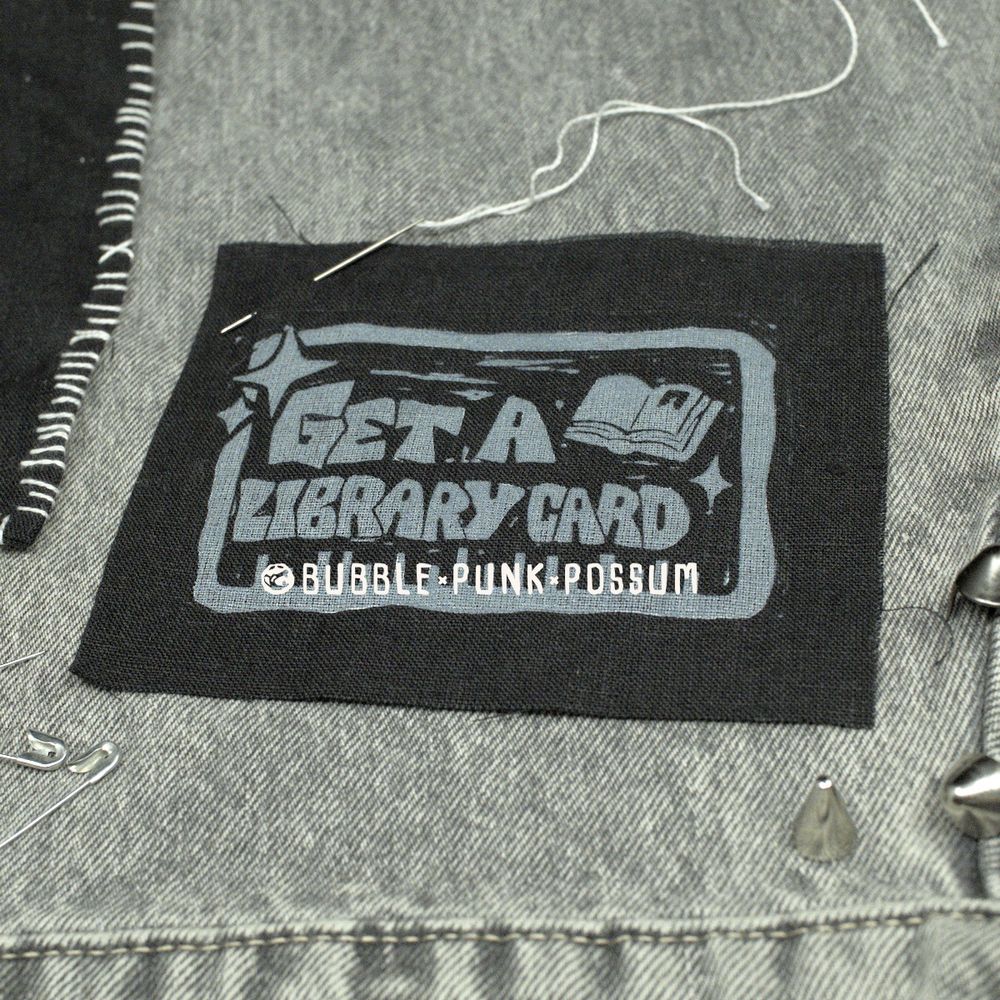 A black patch on top of gray denim. The patch design features a rectangular border and the phrase "Get a Library Card" with an open book in the top right corner and sparkles in the top left corner. 
