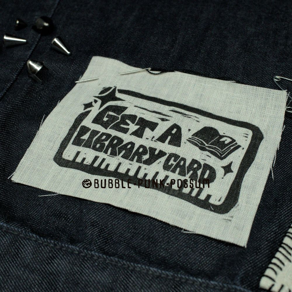 A white patch stacked on top of dark denim. The patch design features a rectangular border and the phrase "Get a Library Card" with an open book in the top right corner and sparkles in the top left corner. 