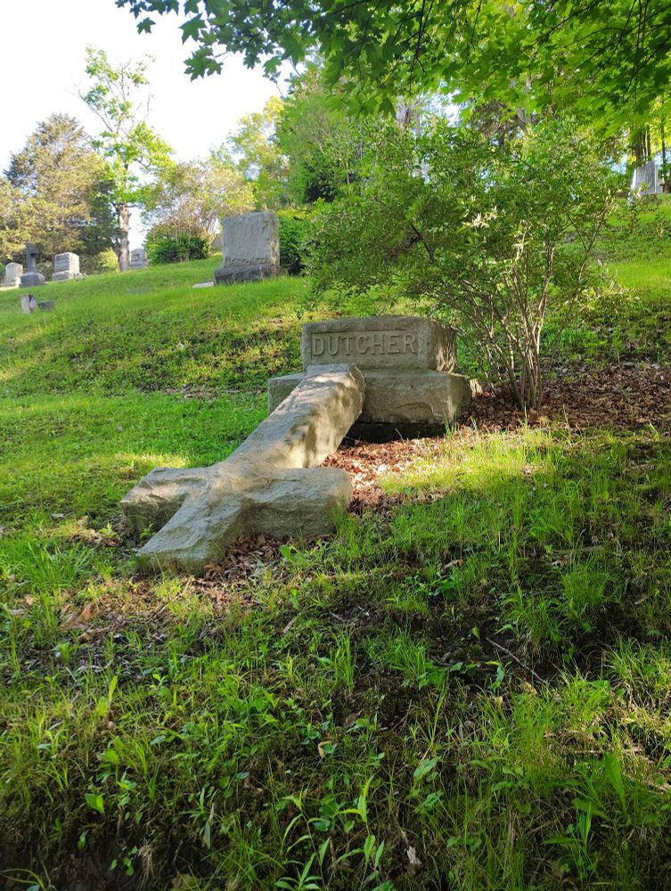 Photo of a broken gravestone where a large stone cross has fallen forward on the ground.