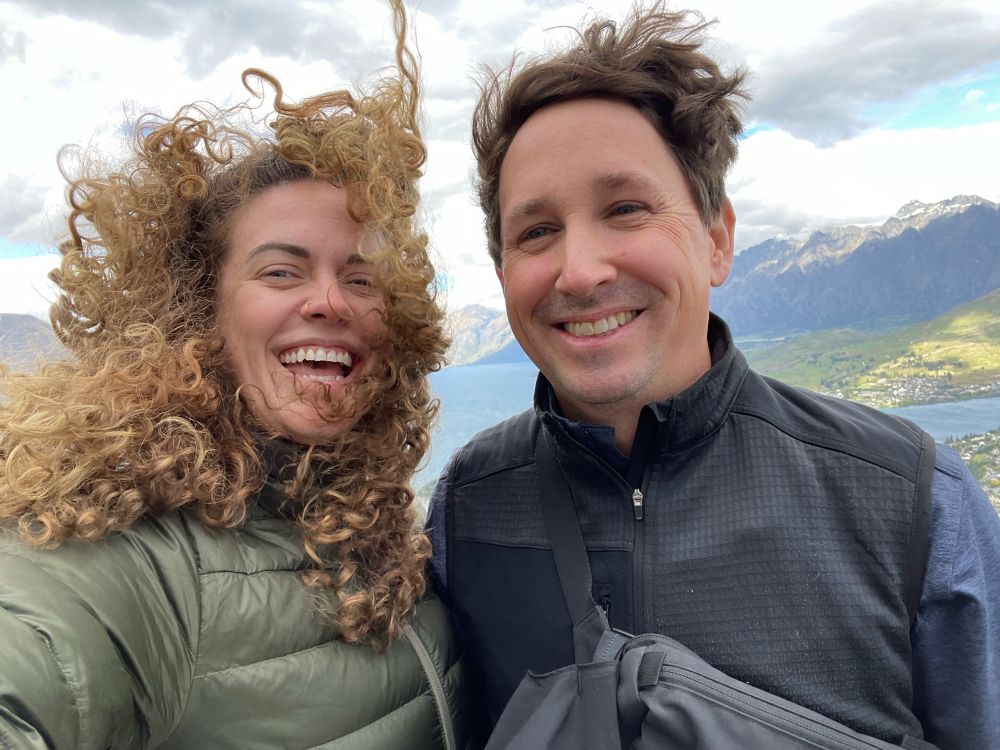 At the top of the lift in Queenstown NZ, wind whipping my curly hair into my face with a beautiful view of snow capped mountains and water behind us 