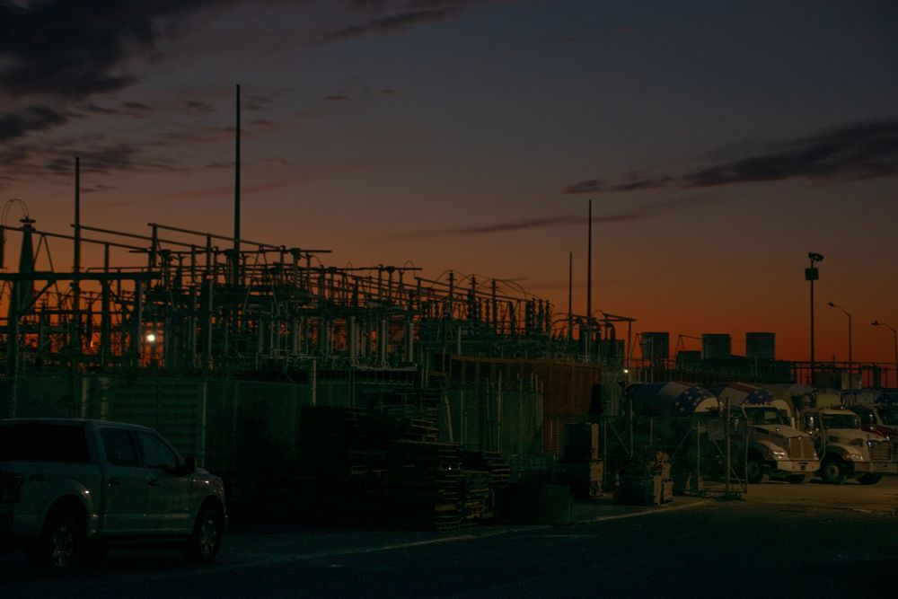 Electrical plant and trucks down by the bay at dusk with an orange glow at the horizon. 