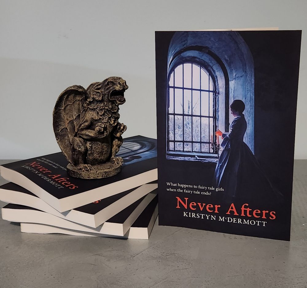 Copy of the collection Never Afters by Kirstyn McDermott standing alongside a small stack of additional copies. The cover features a woman in historical garb standing before an arched window looking out on a wintry landscape. Sitting on the stack of books is an enthusiastic-looking gargoyle. Or maybe he's just hungry ...