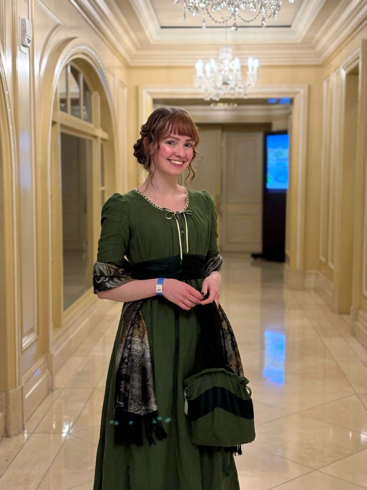 A young woman with auburn hair and bangs smiles at the camera in a cream-colored paneled hallway. She is wearing a green cotton voile regency dress with a green shot silk taffeta sash, a silver and black cashmere wrap draped around her arms, and holding a reticule made out of the same fabrics as her dress.