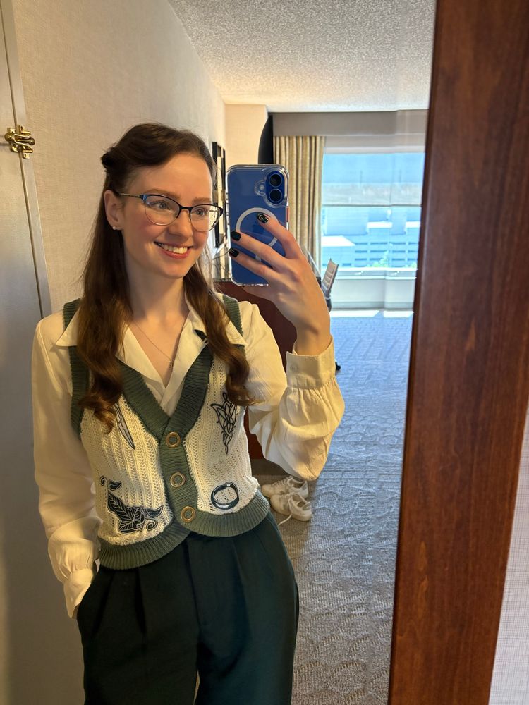 Mirror selfie of a woman wearing a 1930s/1940s cream button down blouse with pleated dark green pants and a chunky knit cream and green Lord of the Rings sweater vest, with green and cream embroidery of the one ring, Sting, the Fellowship of the Ring leaf, and the Evenstar. The woman is also wearing glasses and has her hair twisted back and pinned.