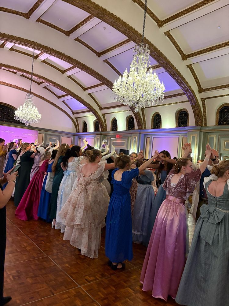 Women in colorful regency dresses and other fancy dresses line up with their hands braced against their partners’ to form a tunnel through which dancers run. They are in the middle of an ornate ballroom with paneled pale green and cream walls, crystal chandeliers, and white and gold ceilings.