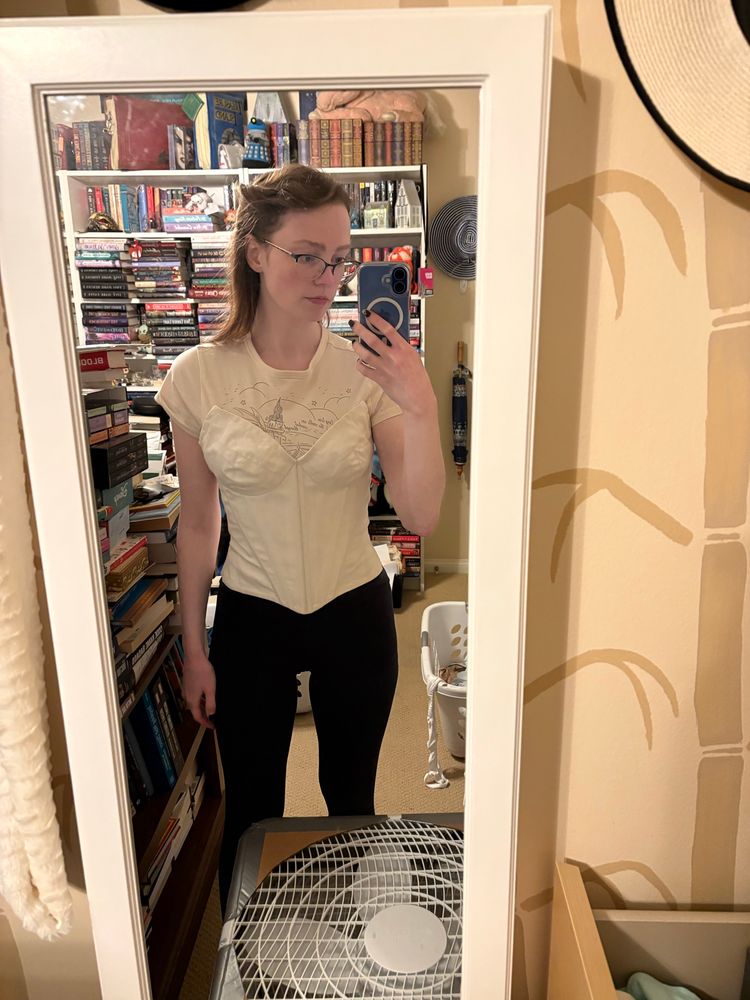 Mirror selfie of a woman wearing a modern? corset over a t-shirt and leggings. The corset is wrinkled around the hips and the cups of the corset are far too large and wrinkling 