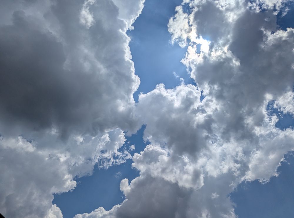 Sunlit clouds in a hazy blue sky. Sun is near upper right. Some of the clouds are dark gray but bright around the edges.