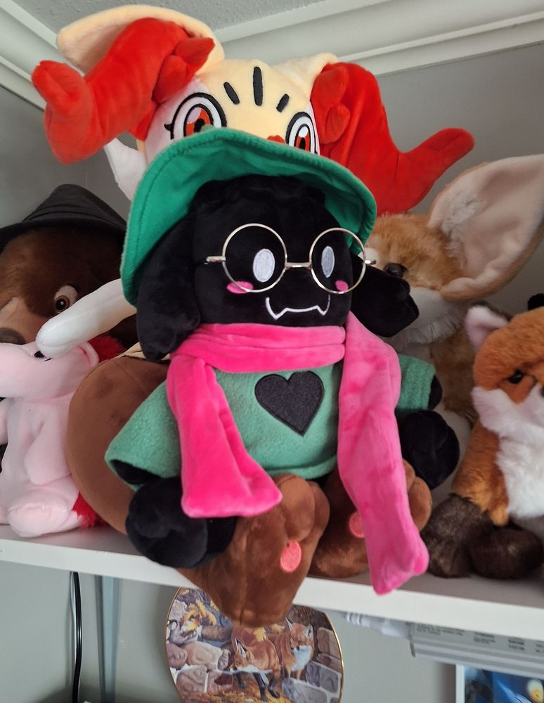 a plush of Ralsei from DELTARUNE on the lap of a larger braixen plush, which is itself positioned on a shelf with other plushies.