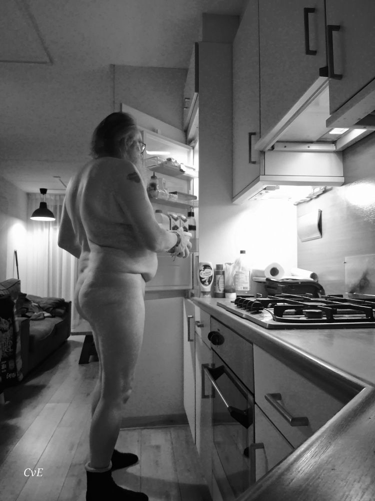 A nude man is standing in front of an open fridge. He takes something from it. On the right is a stove and an oven, on the left a jacket hangs from a chair.