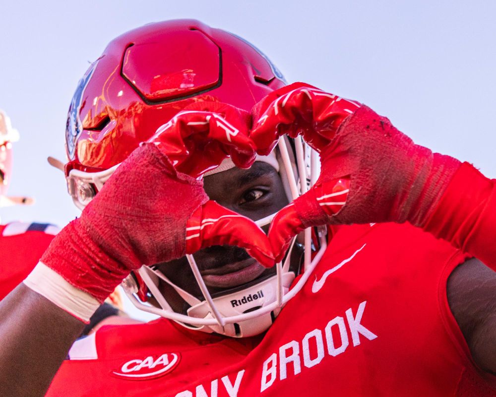 A Stony Brook football player in a red uniform making a heart shape with their hands in front of their face.