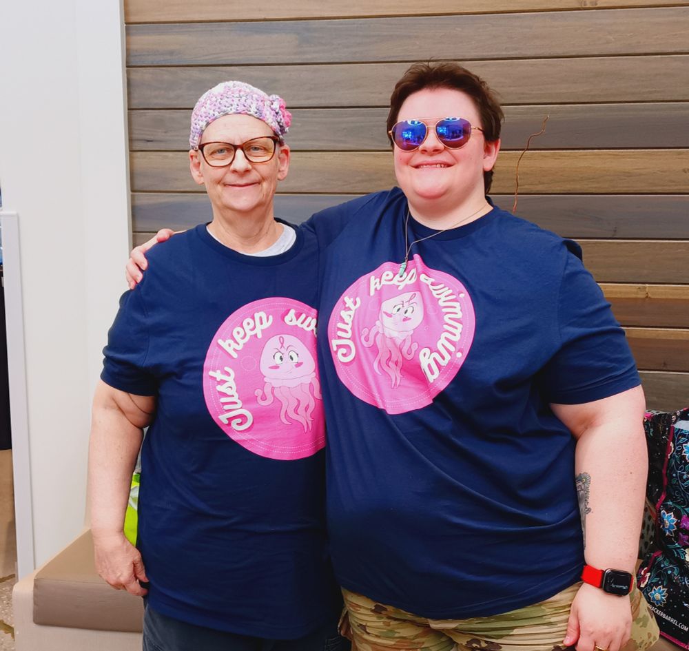 Photograph of two women in matching blue shirts with the design from the previous image. The left woman is older, with glasses and a knit cap. The right woman is younger, slightly taller, with short brown hair and aviator sunglasses.