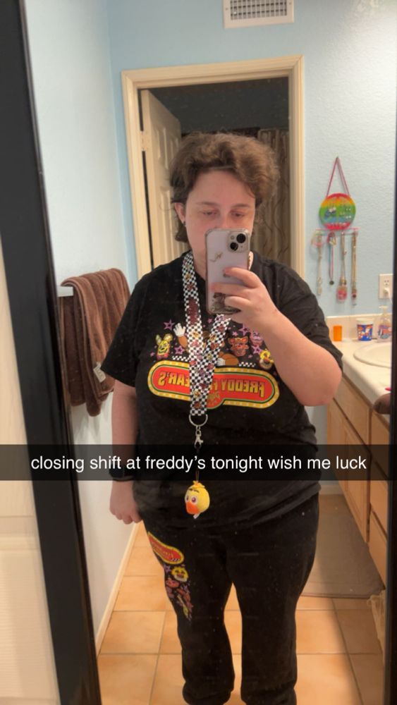snapchat mirror selfie. i’m wearing a fnaf t-shirt, fnaf sweatpants, and a fnaf lanyard with a chica plushie keychain hanging from it. the snapchat caption reads “closing shift at freddy’s tonight wish me luck”
