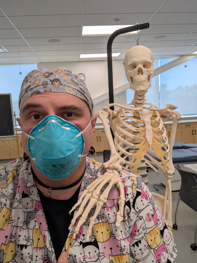 A medical student wearing a green n95 mask, a Studio Ghibli scrub cap, and cat scrubs with a skeleton with its hand on the med students shoulder
