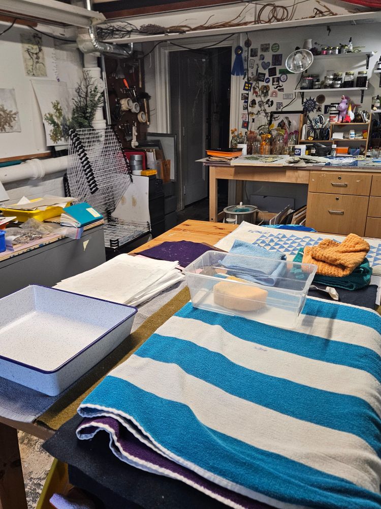A well-used artist's studio prepared for papermaking. In the foreground, several colorful towels, felts, and white paper sheets are stacked on a large wooden table alongside a white enamel basin and a plastic tub with sponges. The background reveals a busy workspace with shelves of jars, tools, art supplies, and pressed plants, along with drying racks and artwork pinned to the walls. 

