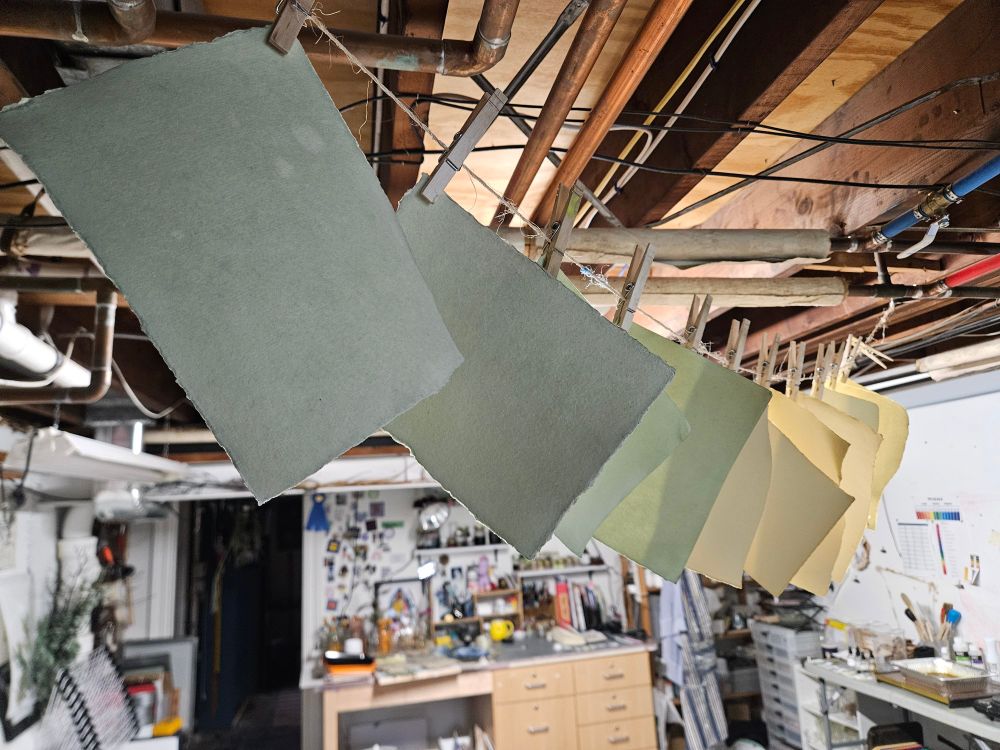 Dyed handmade papers in shades of green and yellow hang from a clothesline with clothespins in Allison's basement studio. The papers were naturally dyed using violets. The background shows shelves, cabinets, art supplies, and pinned inspiration images, contributing to a creative workspace atmosphere.

