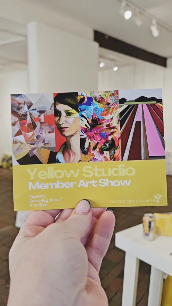 Alt text: A hand holds a colorful postcard in a white-walled art gallery. The postcard promotes the “Yellow Studio Member Art Show,” featuring four vertical art samples: a geometric abstract, a stylized portrait, a vibrant floral composition, and a graphic landscape with fields. Text on the yellow portion of the postcard reads: “Yellow Studio Member Art Show. Opening Saturday June 7, 4 to 6pm. On view June 7 to July 5.” A table with a can and yellow materials is visible in the background.

