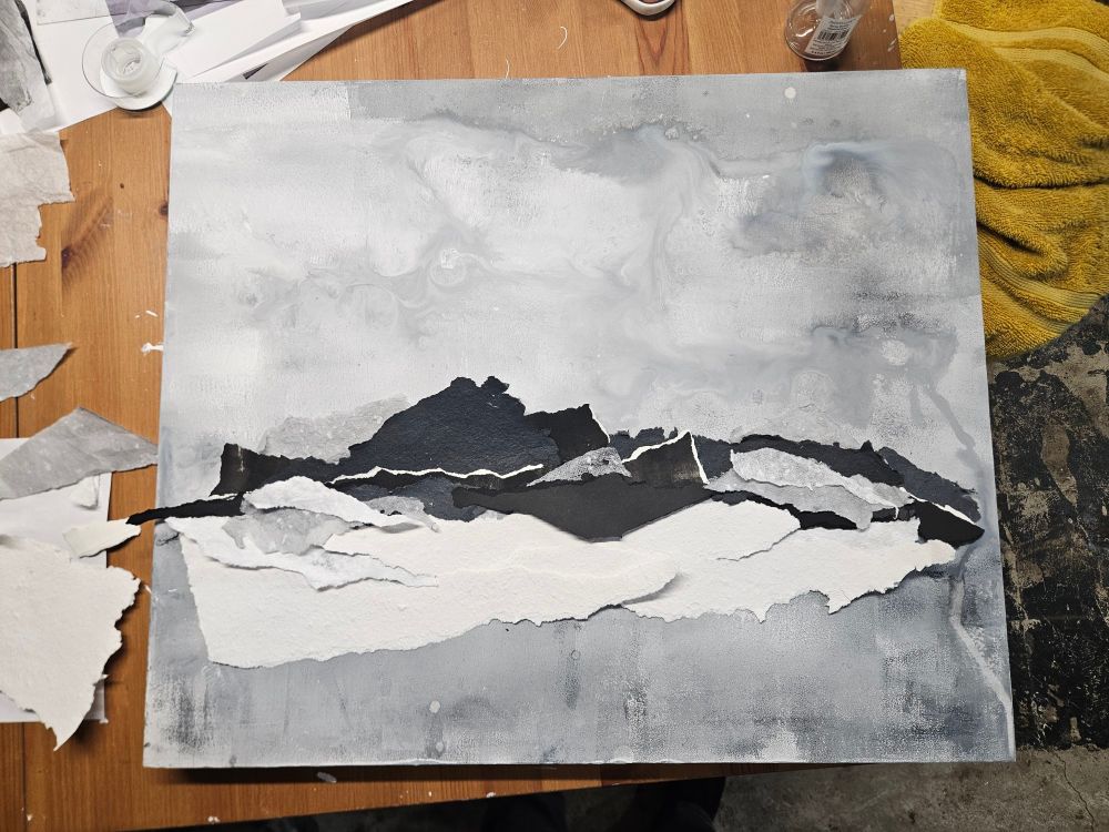 An in-progress collage artwork resting on a wooden table. The background features a cloudy, marbled blend of gray and white paint, evoking a moody sky. Torn pieces of paper in shades of black, gray, and white are layered horizontally across the center, resembling a mountainous landscape or horizon line. Scraps of torn paper, tape, and tools surround the collage, indicating an active workspace.