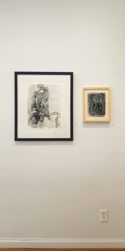 A gallery wall at Upstream Gallery in Hastings-on-Hudson, NY, displaying two framed artworks. On the left is a larger black-and-white abstract drawing of a seated human figure, framed in black. To the right is a smaller work in a light wood frame, also monochrome, featuring overlapping white contour lines of figures on a dark background. Both artworks are hung side by side against a white wall.
