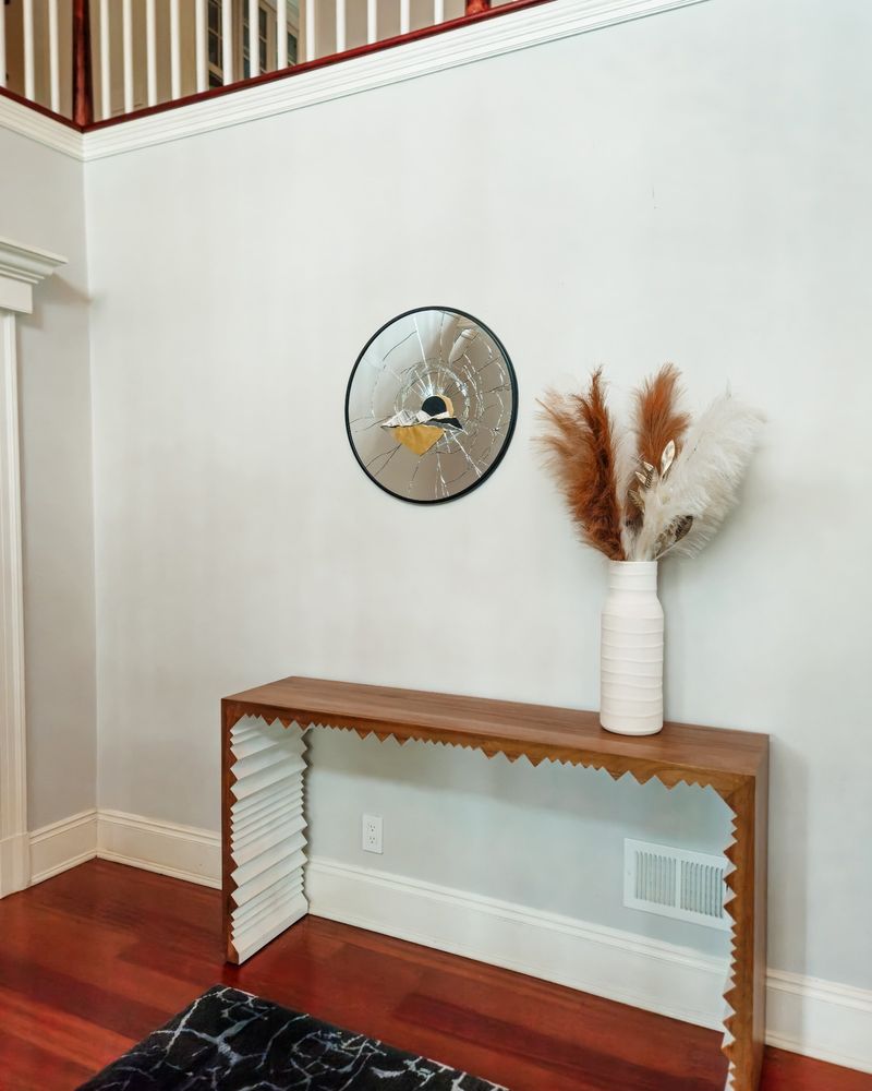 A modern entryway with a circular, abstract mirror artwork featuring cracked glass and layered geometric elements in black, white, and gold tones. The mirror is mounted above a sculptural wooden console table with jagged, zig-zag side panels. A tall white vase with pampas grass in earthy tones sits atop the console. 


