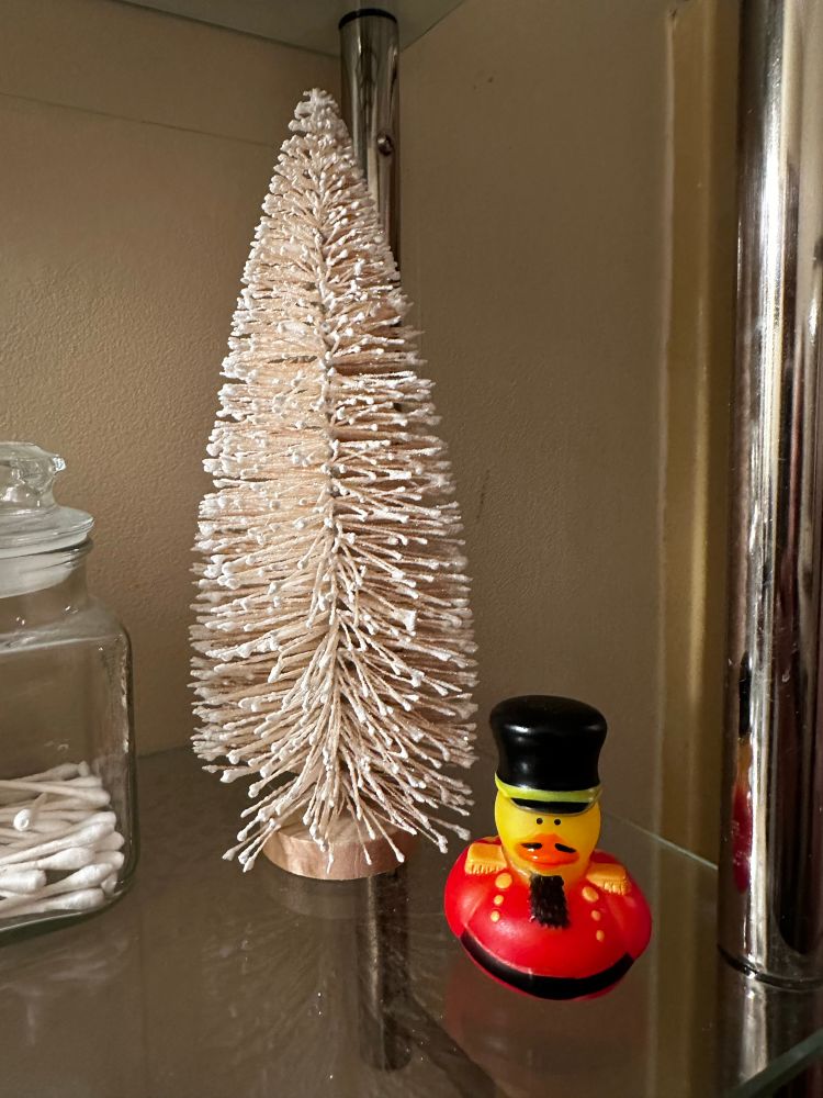 A pink-white bottle brush tree next to a small nutcracker rubber duck on a glass shelf