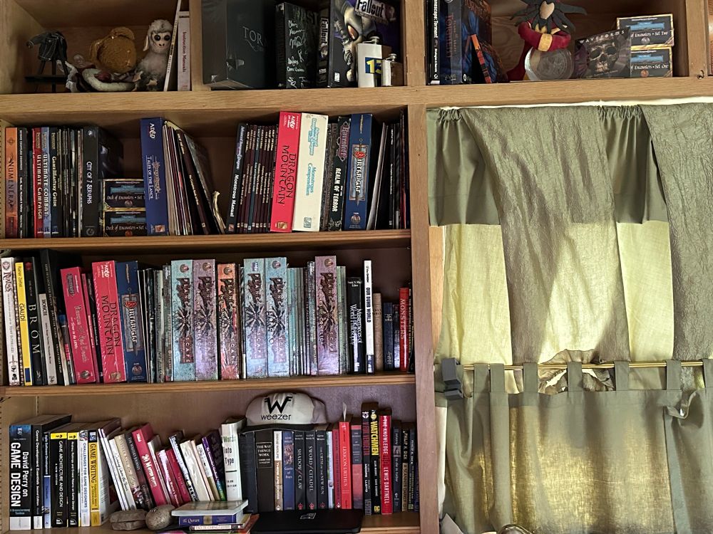 A shelf of books, boxed sets, and accessories for various TTRPGs. Also a Weezer tour hat from '95 and a Lady of Pain plush.