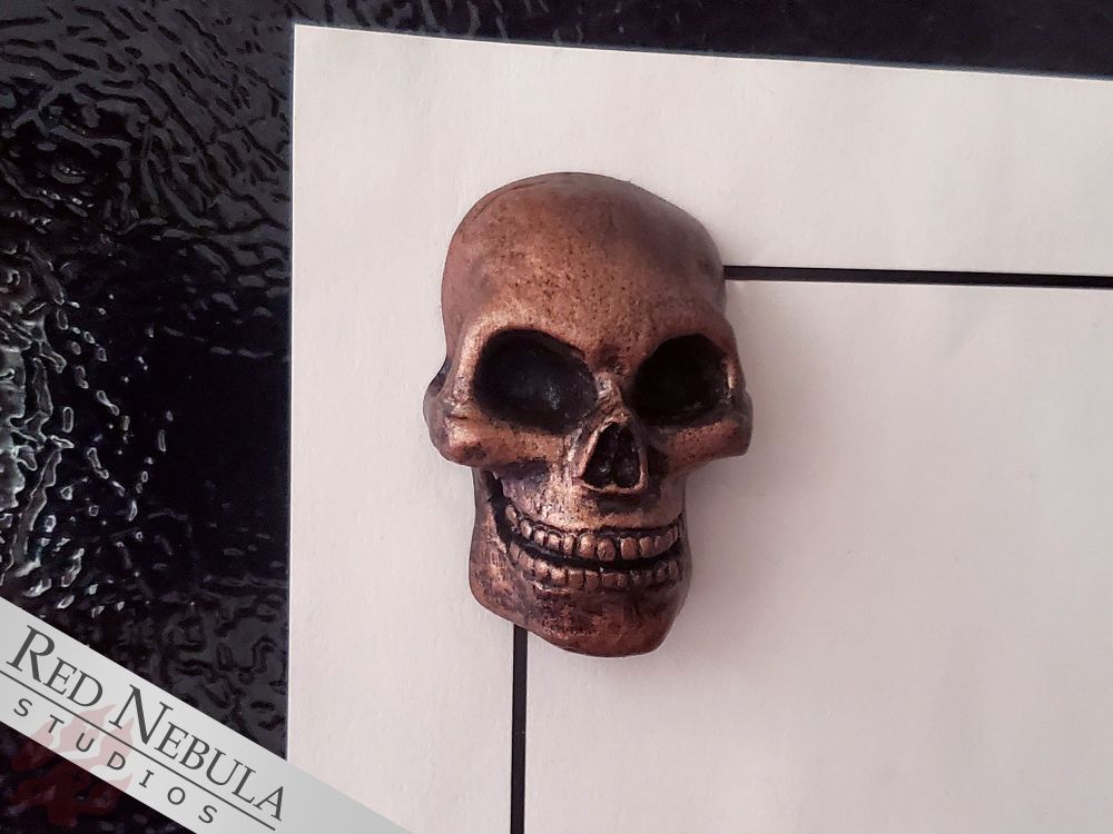 A flat-back resin human skull with an embedded magnet holding a piece of paper to a refrigerator. The skull is painted to look like aged bronze.