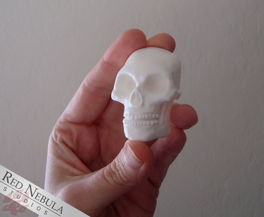 A hand holding a small, white human skull made of cast resin. It is flat on the back.