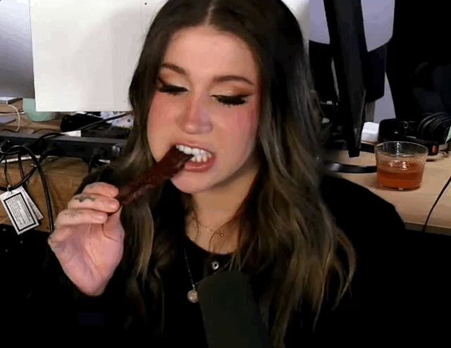 a woman is eating a piece of meat in front of a computer monitor and a glass of liquid