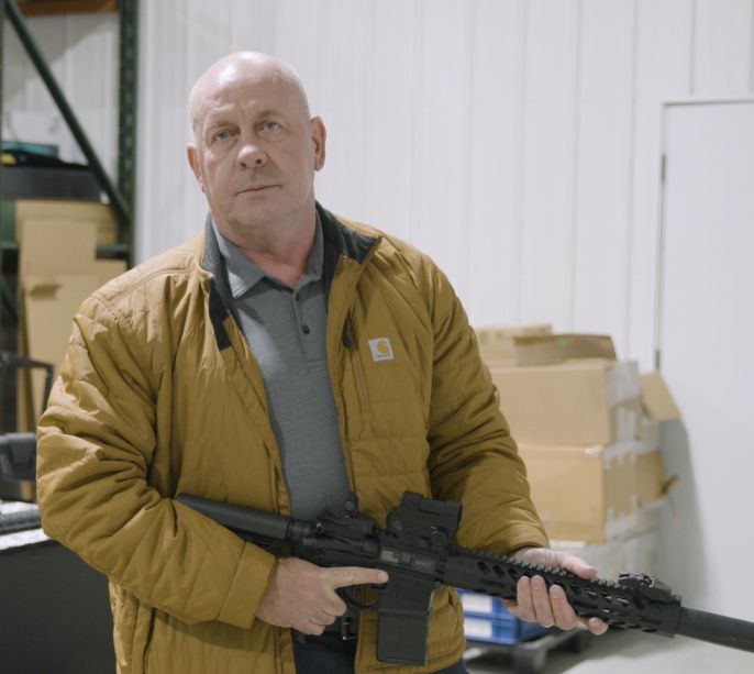 Image taken from "Dave Taylor for Congress" website, showing Taylor standing in a warehouse and holding a semiautomatic rifle. https://davetaylorforcongress.com/meet-dave/
