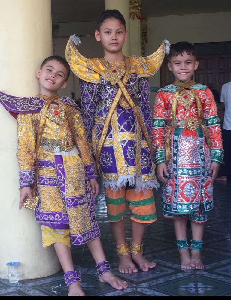 my 3 sons (aged 10, 7 and 7) in traditional Thai costumes worn to perform Khon dances.  Eldest  in the center and 2 on his right wear gold and purple ‘giants’ outfits with ornate, outsize, shoulder ‘armor’. 3 on the other side of big brother is in a red and green monkey outfit, basically the same style but no shoulder armor.  All have cute matching ankle cuffs and extreme amounts of costume jewelry.  They look happy and relaxed but didn’t much enjoy it at the time, who wants to get dressed up in this heat!
