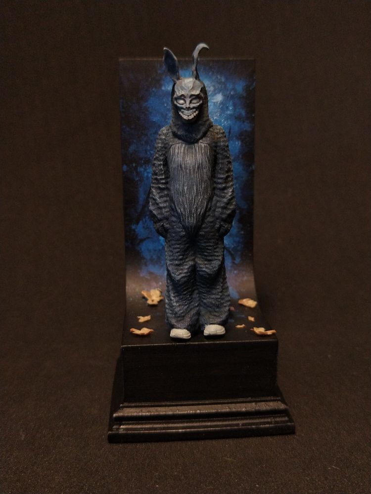 Das Bild zeigt eine detaillierte Figur in einem bunten, tierähnlichen Kostüm, die auf einem schwarzen Podest steht. Die Figur hat lange Ohren und ein maskenhaftes Gesicht mit einem gruseligen Lächeln. Sie trägt eine dunkelgraue, rissige Textur, die an Fell erinnert. Die Figur steht auf einem schwarzen Podest, das mit einigen gelben Blättern verziert ist. Im Hintergrund ist eine künstlerische Darstellung eines blauen Nachthimmels mit dunklen Ästen zu sehen. Die Figur ist in einer ruhigen, aber mysteriösen Pose dargestellt, was durch die dunkle Umgebung und die künstlerische Hintergrundgestaltung verstärkt wird.