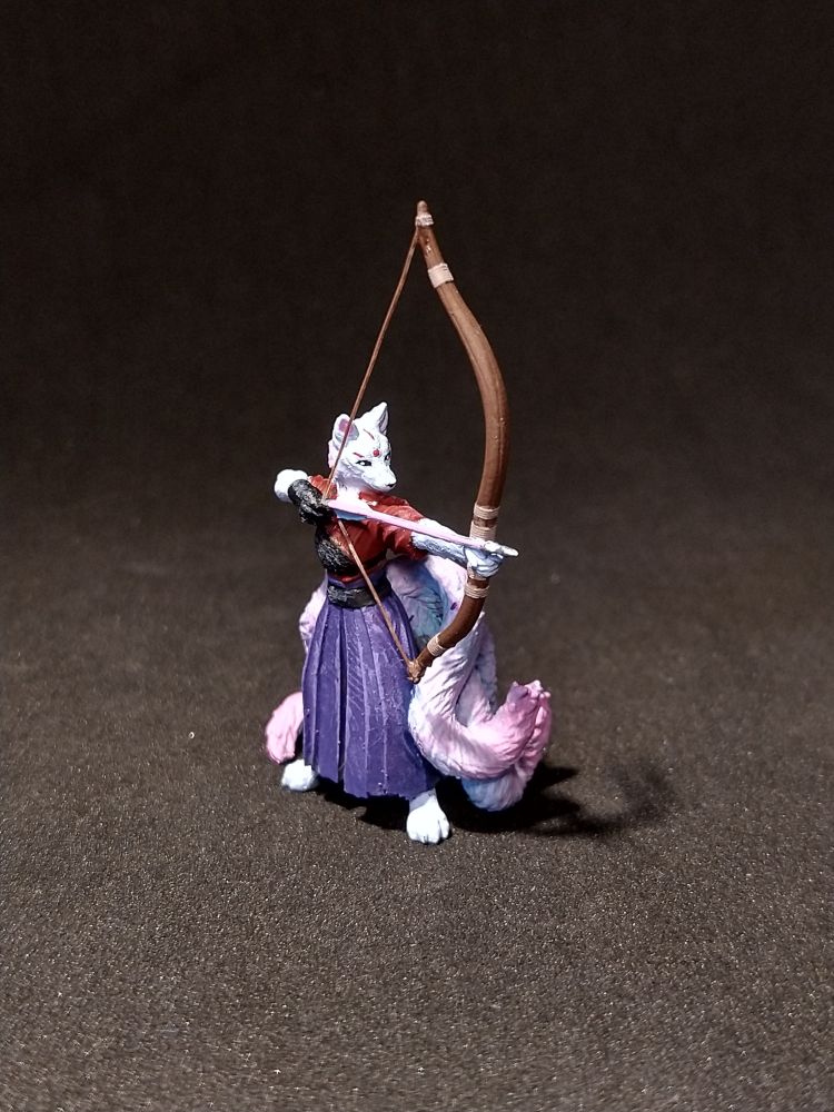 Das Bild zeigt eine detaillierte Miniaturfigur eines Wesens, das wie ein Wolf aussieht, das einen Bogen spannt. Das Wesen hat eine weiße Felle und trägt ein rotes Oberteil mit einem schwarzen Gürtel und einen langen, violetten Rock. Der Bogen ist aus Holz und hat eine geschwungene Form, die von den Händen des Wesens gehalten wird. Der Schaft des Bogens ist dünn und braun, und die Sehne ist sichtbar. Der Hintergrund ist dunkel, was die Figur hervorhebt. Die Figur steht auf einem flachen, dunklen Untergrund, der die Details der Figur betont.