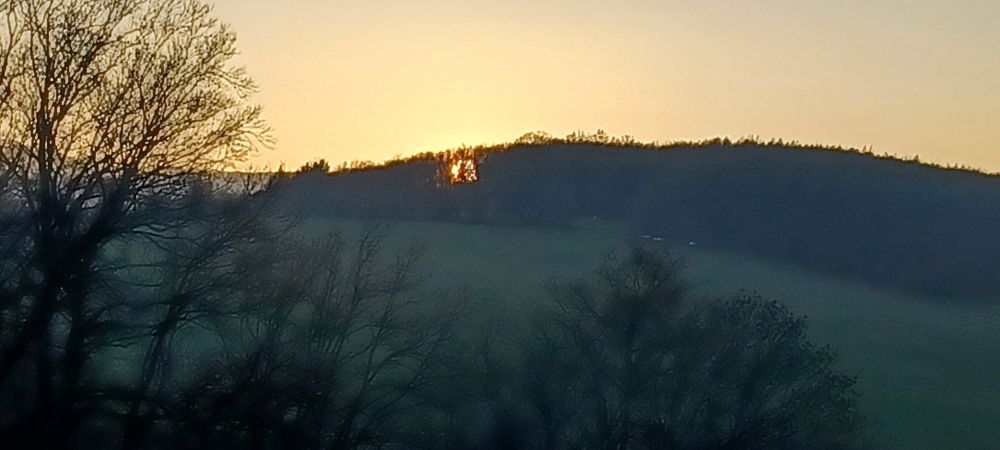 Recht dämmerig, Blick auf Hügel gegenüber, Sonne untergehend zwischen den Bäumen des Hügels