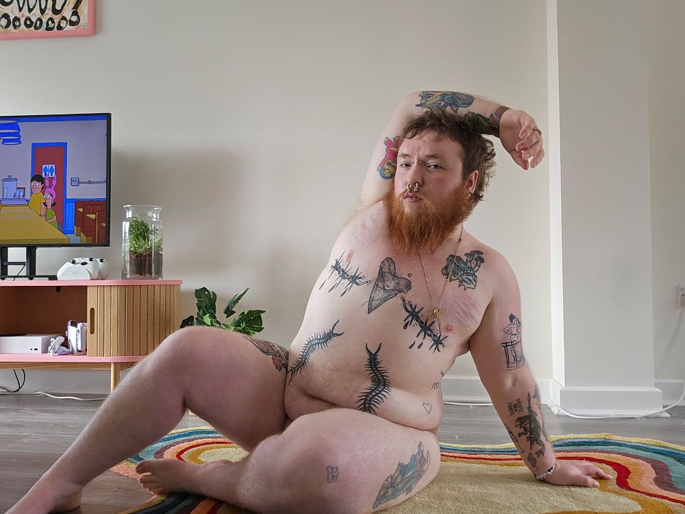 A photo of me sitting on a rug in my living room, one leg is bent and one arm is over my head. I am tattooed on most available skin. My hair is brown and messy and I have a big ginger beard 
