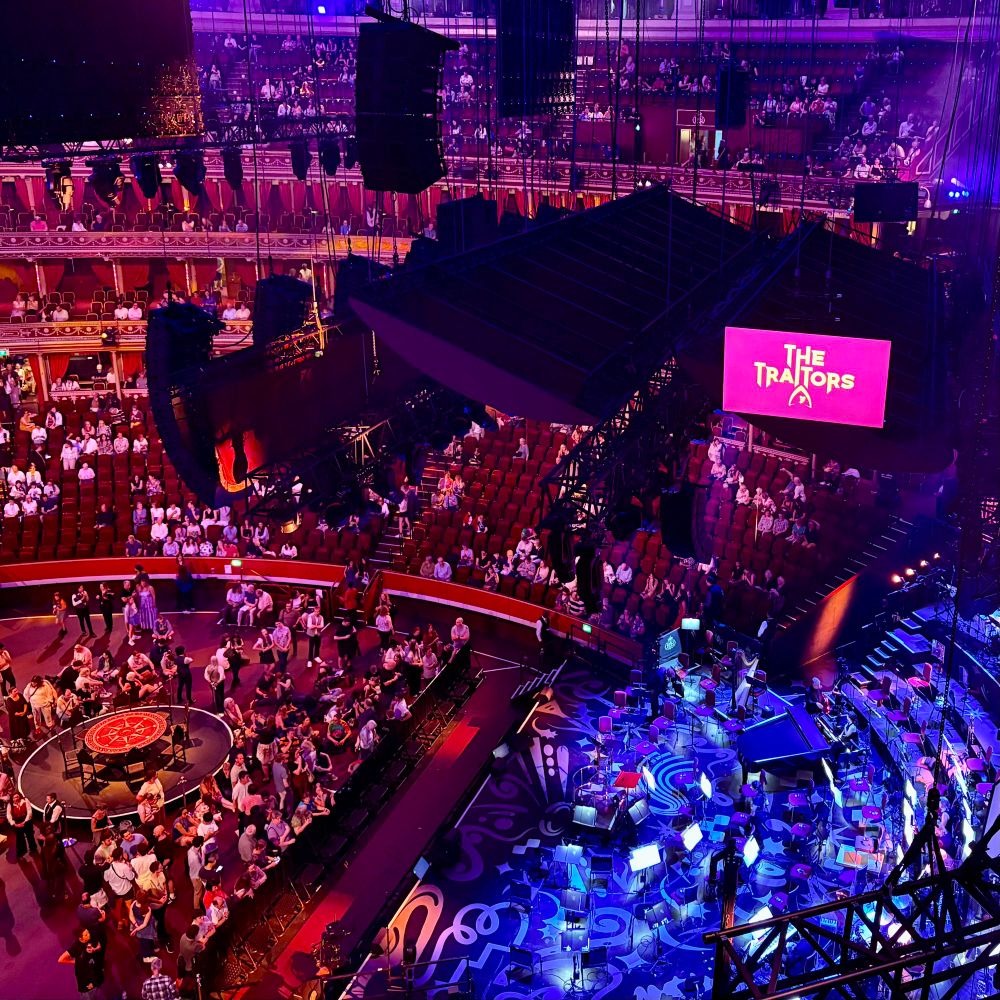 A crowd of music-lovers bathed in purple and blue light. The Traitors logo is in the background: who do you trust?