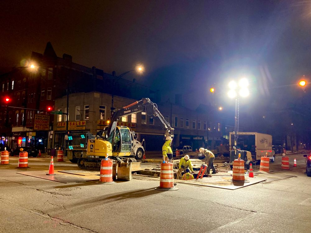 Construction digging into the middle of the intersection at Ashland and Chicago avenues, lights on at night.