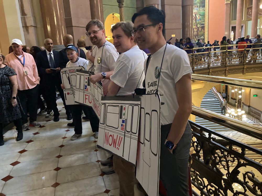 A group of transit activists wear a cardboard CTA train that reads “Fund Transit Now!” In the Springfield Capitol Rotunda.