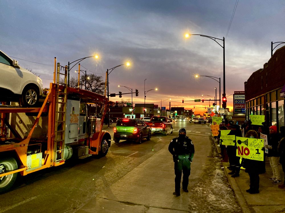 A sign saying “No to 1 lane” in a crowd of protestors for and against, as a CPD officer walks on the road, and a multi car tow truck waits at the light, at Archer and Pershing.