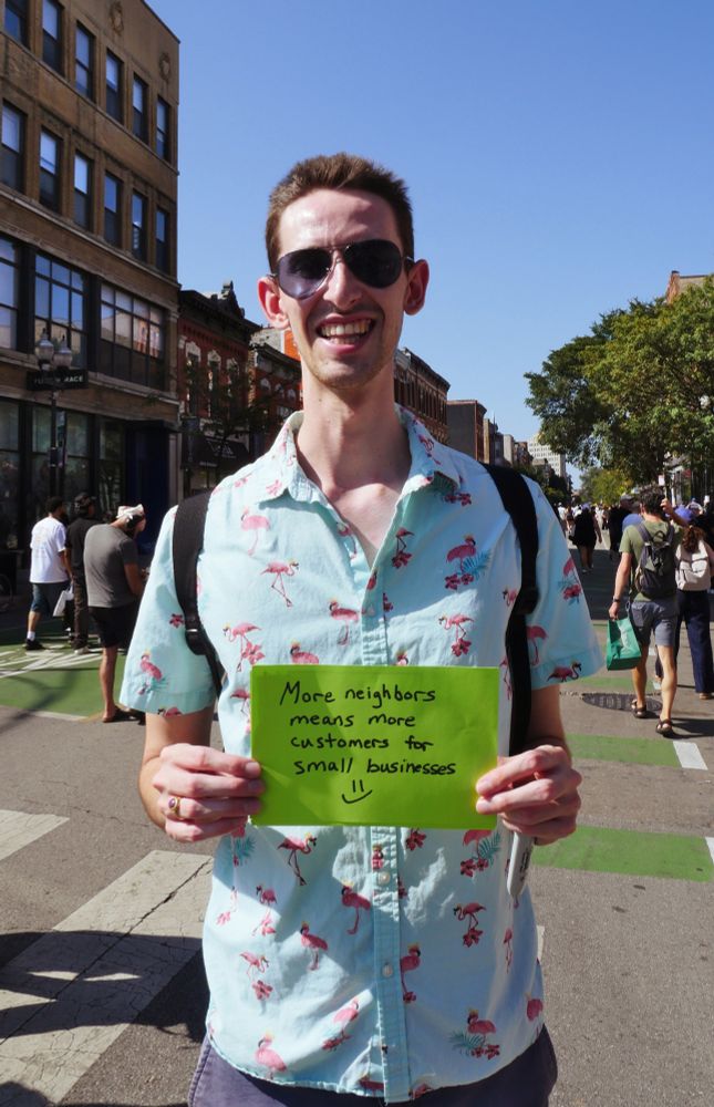 Robert in a flamingo shirt holds a sign reading "More neighbors means more customers for small business (and a smiley face)"