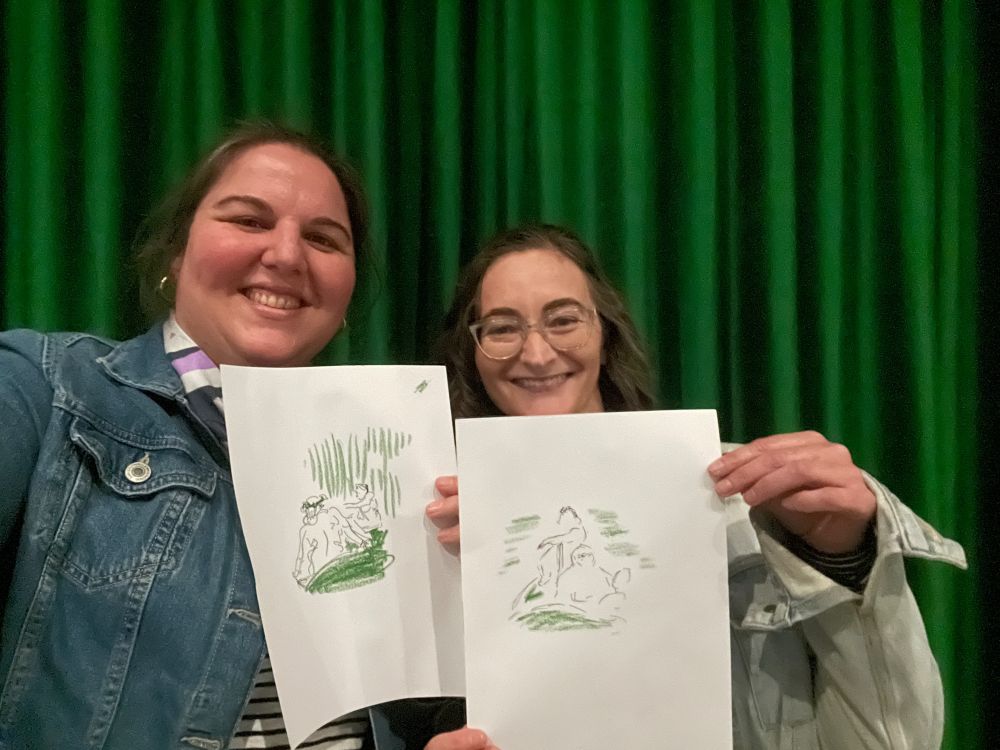 Me and Jo, in front of a green velvet curtain, both wearing denim jackets, holding our sketches in black pen and green pastel, each of two figures lounging 
