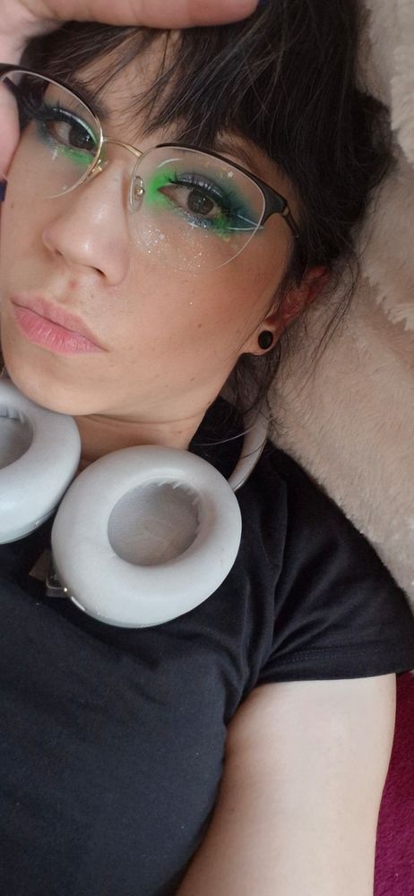 Selfie von mir auf dem Sofa liegend. Mit zerzaustem Haar, Kopfhörer um den Hals, blau grün lilanes Make up mit jeder Menge Glitzer und weißem Eyeliner und false Lashes.
