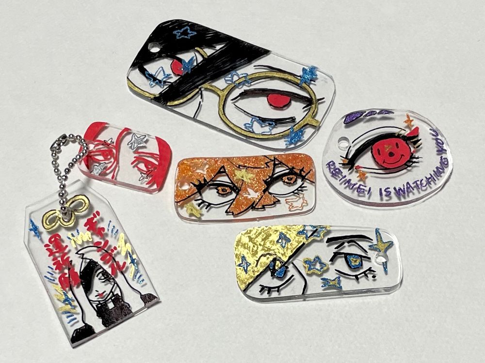 プラ板で作ったキーホルダーの写真。キャラクターの目元のアップが描かれたもの、お守りの形のものがある。