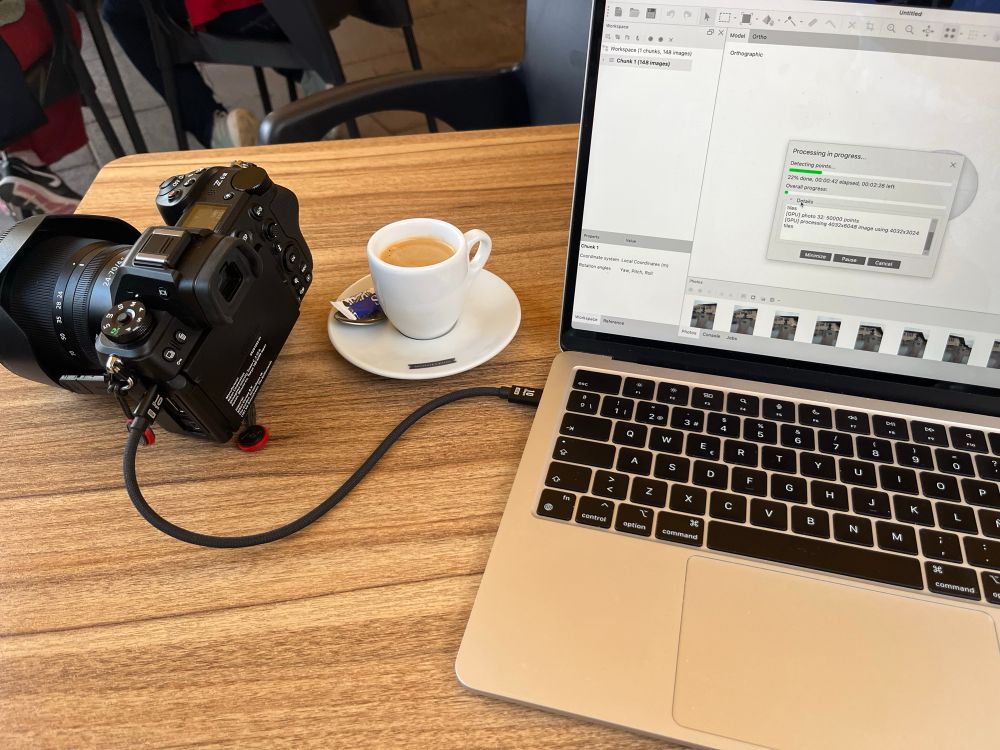Mesa de bar en la que se ve una cámara de fotos conectada por cable a un ordenador portátil que muestra en su pantalla la barra de progreso del programa de fotogrametría, entre ambos una taza de café.