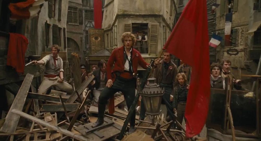 Still from “Les Miserables” in which Enjolras, from the barricade, holds up an enormous red flag.