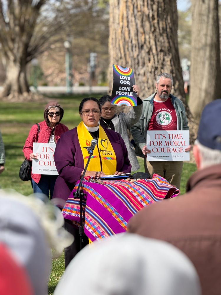 The Rev. Dr. Sofía Betancourt stands at a microphone, speaking during an outdoor rally. She wears dark glasses, a clerical collar, a gold stole that reads “SIDE WITH LOVE,” and a purple coat. In front of her is a colorful textile with bright stripes draped over a table or podium holding papers. Behind her stand several people holding signs. One sign directly to her left reads “LOVE GOD, LOVE PEOPLE” with rainbow arches above and below the text. Two other signs, one to the right of the speaker and one to the left of the speaker, both read “IT’S TIME FOR MORAL COURAGE” with “SOJOURNERS” written beneath. The background features large trees, green grass, and a park-like setting. The focus is on the speaker, with blurred heads and shoulders of attendees visible in the foreground.