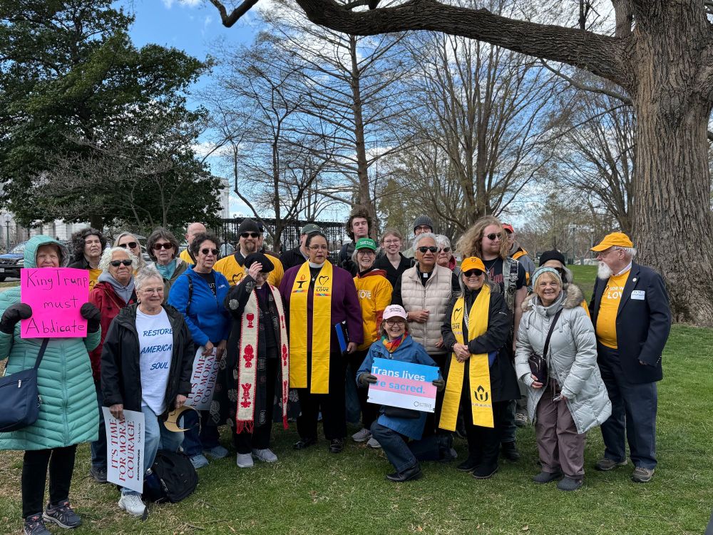 A group of people with The Rev. Dr. Sofia Betancourt in the center pose together outdoors with on a grassy area with trees and a bright blue sky in the background. Several individuals wear bright yellow stoles that say “SIDE WITH LOVE,” indicating affiliation with the Unitarian Universalist faith. Many are also dressed warmly in coats and hats. Some people hold signs with messages such as “King Trump must Abdicate,” “Trans lives are sacred,” “RESTORE AMERICA’S SOUL,” and “IT’S TIME FOR MORAL COURAGE.” 